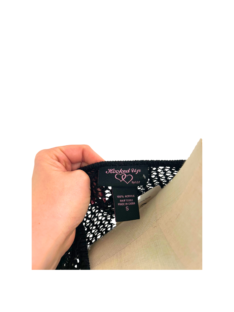 Hand holding a label from a black Crochet Tie Cardigan from the brand Hooked Up on a white background